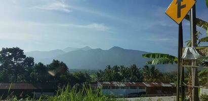 A view of green rice fields and gardens with blue sky and white clouds and the silhouette of a mountain photo