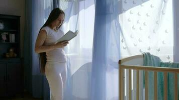 Future young mother between 30 and 35 years old is reading a book close to the open window. video