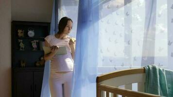 Future young mother between 30 and 35 years old is reading a book close to the open window. video