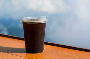 A glass of iced coffee with iced cube puts on table with sky and cloud background. Summer drink concept. photo