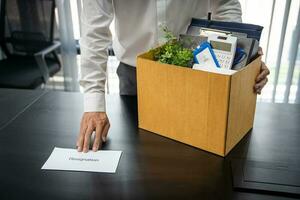 Business man sending resignation letter and packing Stuff Resign Depress or carrying business cardboard box by desk in office. Change of job or fired from company photo