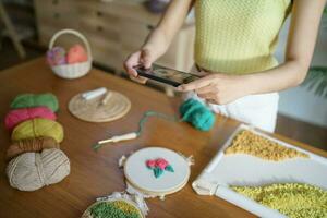 asiático mujer tomando teléfono inteligente foto puñetazo aguja. teléfono destino en social redes en estudio taller. diseñador lugar de trabajo hecho a mano arte proyecto bricolaje bordado