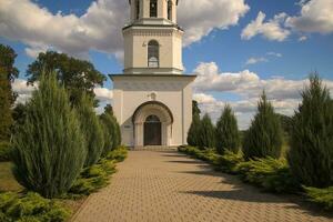 An alley of cypresses leads to an old white chapel with a bell. The shadows of plants indicate the entrance under a canopy, the door with a cross, the facades of the windows. Summer sunny day. photo