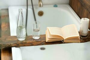 a book, a bottle of water, a glass and a candle on a wooden tray above the bathroom, against the background of an antique faucet photo