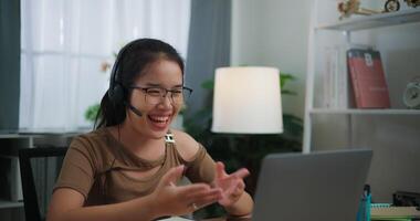 beeldmateriaal van gelukkig jong Aziatisch vrouw vervelend bril en koptelefoon aan het doen video conferentie Aan een laptop Aan een bureau in de leven kamer Bij huis. levensstijl, werkzaamheid en mensen concepten.