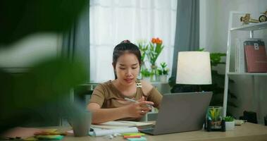 imágenes de joven asiático mujer aprendizaje en línea con ordenador portátil en un escritorio y escritura en papel en el vivo habitación a hogar. estilo de vida, actividad y personas conceptos. video