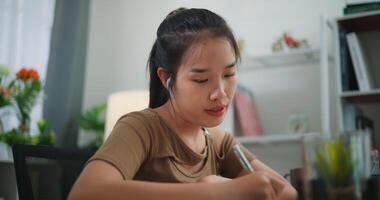 metraggio di giovane asiatico donna apprendimento in linea con il computer portatile su un' scrivania e scrittura su carta nel il vivente camera a casa. stile di vita, attività e persone concetti. video