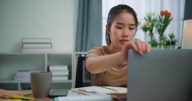 beeldmateriaal van jong Aziatisch vrouw aan het leren online met laptop Aan een bureau en schrijven Aan papier in de leven kamer Bij huis. levensstijl, werkzaamheid en mensen concepten. video