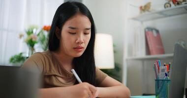 metraggio di giovane asiatico donna apprendimento in linea con il computer portatile su un' scrivania e scrittura su carta nel il vivente camera a casa. stile di vita, attività e persone concetti. video