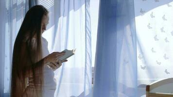futur Jeune mère entre 30 et 35 ans vieux est en train de lire une livre proche à le ouvert la fenêtre. video