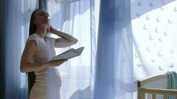 Future young mother between 30 and 35 years old is reading a book close to the open window. video