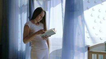 Future young mother between 30 and 35 years old is reading a book close to the open window. video