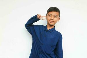 Smiling confident boy wearing a blue casual shirt looking at the camera while showing v sign, isolated on white background photo