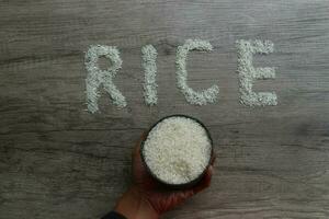 Hand holding an iron container filled with rice, and rice grains forming the letters RICE on a wooden background photo