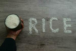 Hand holding an iron container filled with rice, and rice grains forming the letters RICE on a wooden background photo