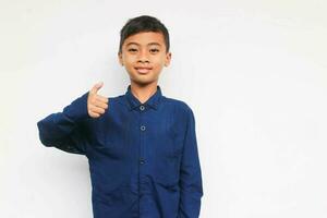 Smiling confident boy wearing a blue casual shirt looking at the camera with showing thumb up isolated on white background photo