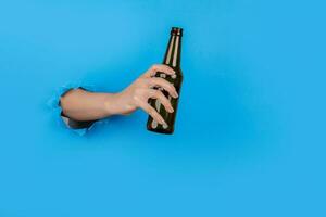 Hand through paper holding alcohol bottle on blue background photo