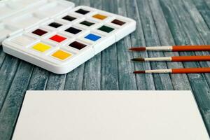 A set of new and unused watercolors, brushes and a piece of white paper on a textured surface ready to be used photo