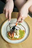 Toast with salmon and a poached egg on an avocado spread. Female hands with a fork photo