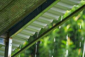 Raindrops on a metal roof gutter closeup against a blurred background of green trees photo