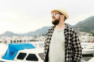 hermoso hombre vistiendo sombrero y lentes cerca centro de deportes acuáticos con yates retrato de riendo hombre con mar Puerto antecedentes con Copiar espacio foto