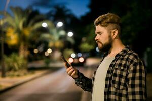 Bearded man holding scrolling texting in his cellphone at night street. Guy calls for taxi in an app in evening city - copy space and place for advertising photo