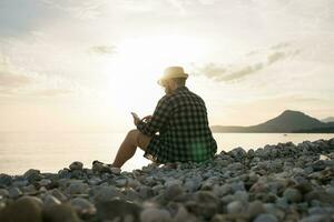 Rear side view of man with smartphone sitting on beach terrace on sunny day outside background with copy space - vacation or freelance or social network concept photo