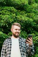 Portrait of attractive cheerful guy using smartphone for scrolling on social media over green tree background photo