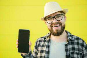 Happy smiling bearded wearing hat man showing mobile phone on yellow background - empty space and place for advertising photo