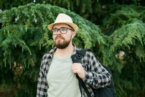 Handsome guy tourist with backpack wearing straw hat for travelling, standing against green tree background outdoors. photo