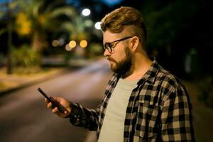 Bearded man wearing eyeglasses is holding scrolling texting in his cellphone at night street. Guy calls for taxi in an app in evening city - copy space and place for advertising photo