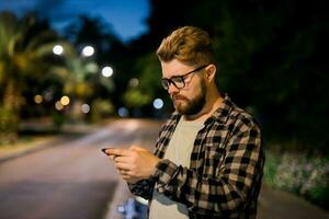 Bearded man wearing eyeglasses is holding scrolling texting in his cellphone at night street. Guy calls for taxi in an app in evening city - copy space and place for advertising photo