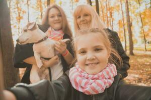 Mother, grandmother and little granddaughter with jack russell terrier dog taking selfie by smartphone outdoors in autumn nature. Family, pets and generation concept photo
