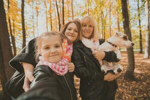 Mother, grandmother and little granddaughter with jack russell terrier dog taking selfie by smartphone outdoors in autumn nature. Family, pets and generation concept photo
