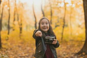 sorprendido niño niña utilizando un Anticuado cámara en otoño naturaleza. fotógrafo, otoño temporada y emociones concepto. foto