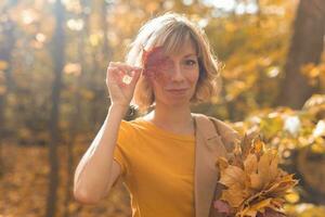 joven rubia mujer cubierta uno ojo con rojo arce hoja. otoño y temporada concepto. al aire libre otoño hembra retrato de cerca con follaje foto
