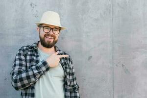 Handsome guy tourist looking happy wearing straw hat for travelling and pointing on empty space, standing against concrete wall background with copy space photo