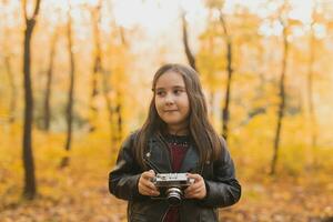 un pequeño niña tomar un foto con antiguo retro cámara en otoño naturaleza. ocio y pasatiempo concepto.