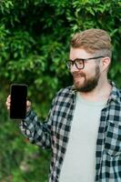 Happy bearded man showing blank smartphone screen over green leaves background - empty space for advertising and copy space photo