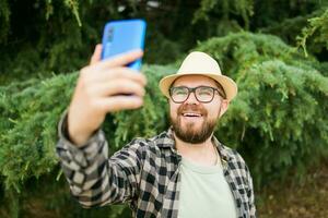 Man taking selfie portrait over tree background - Happy millennial guy enjoying summer holidays in city - Youth and technologies photo