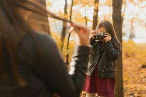 Child girl photographer takes pictures of a mother in the park in autumn. Hobbies, photo art and leisure concept.