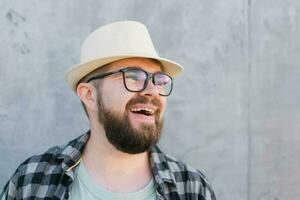 Portrait of positive young hipster man smiling over yellow wall store outdoor background - Handsome trendy bearded guy with hat and glasses standing outdoors - Fashion and trendy youth people concept photo