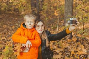 hijo y madre son tomando selfie en cámara en otoño parque. soltero padre, ocio y otoño temporada concepto. foto