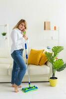 Smiling millennial woman housewife with mop for washes floor enjoys cleaning in minimalist interior. Household cleaning services and domestic work housekeeping photo