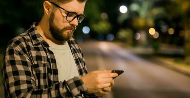 Banner bearded man wearing eyeglasses is holding scrolling texting in his cellphone at night street. Guy calls for taxi in an app in evening city - copy space and place for advertising photo