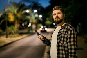 Bearded man holding scrolling texting in his cellphone at night street. Guy calls for taxi in an app in evening city - copy space and place for advertising photo