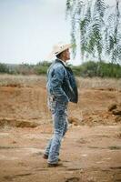 vaquero vistiendo pantalones y un sombrero, en pie debajo el cielo con mullido nubes, trabajando en un granja foto