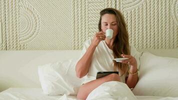 Girl in bed, looking at the tablet and drinking coffee. video