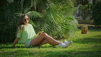 A girl sits in a park on the grass in the shade of palm trees. video