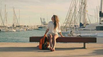 The girl enjoys the sun sitting on a bench in front of the yacht club. video